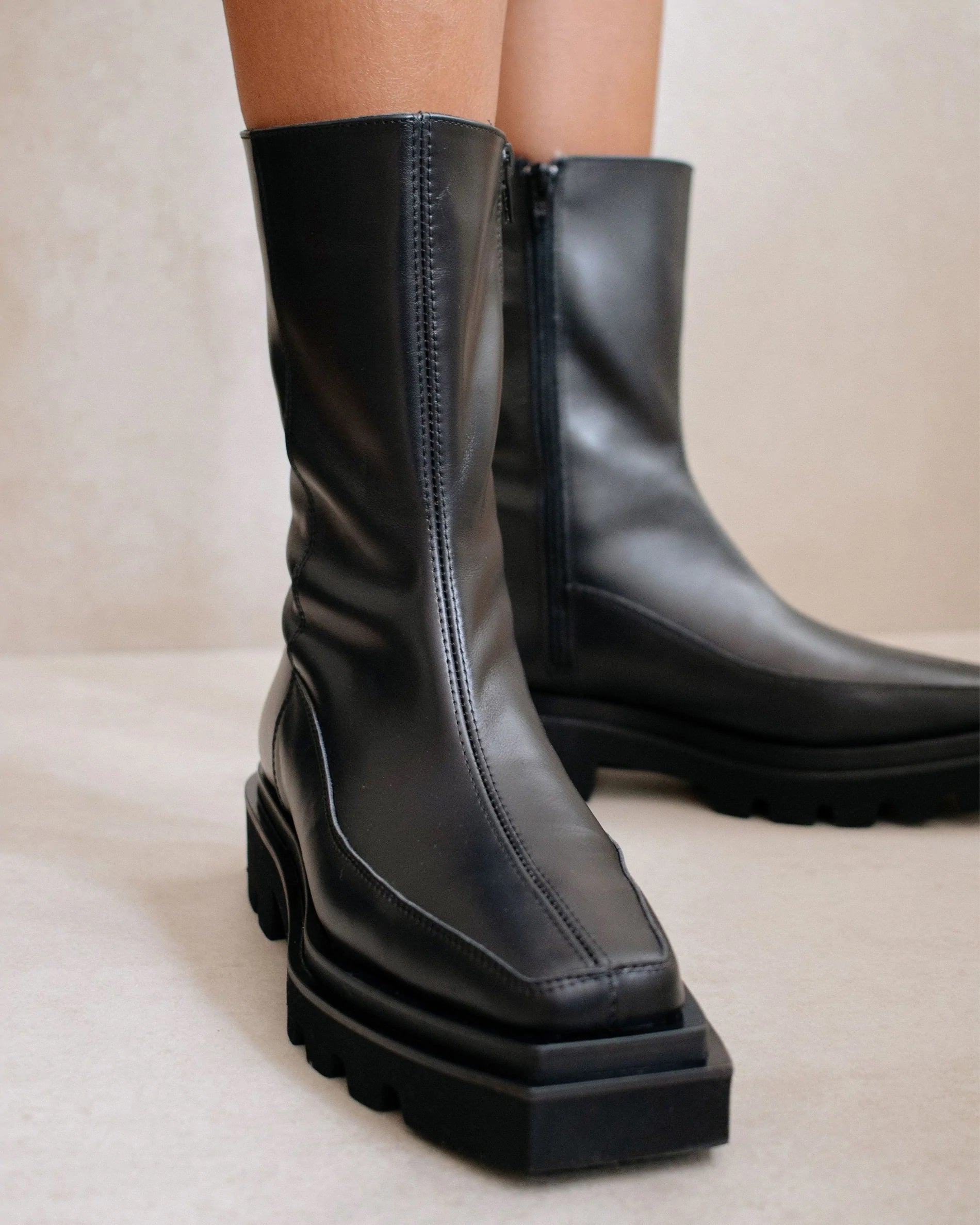 Close-up of the ALOHAS BLOCK TOTAL BOOTS in black, featuring chunky ankle-length design and sturdy resistant soles. These mid-calf boots have slim square toes, visible side stitching, and a partially visible side zipper on the inner sides. The background is neutral and minimalistic.