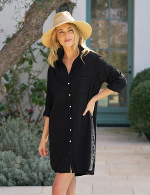 A woman in the FRANK & EILEEN Mary Classic Shirtdress in Famous Denim Black stands outdoors on a stone path, smiling with one hand on her hip. She wears a wide-brimmed straw hat; green plants and a large window are in the background.