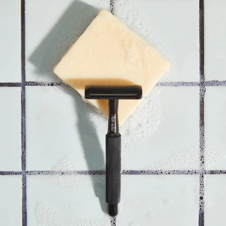 Labeled "KITSCH - SHAVE BUTTER," a black box rests on a cream soap bar with rosemary, shea nuts, and coconut, promoting sustainability and hydration. Features: "Reduces Single-Use Plastic," "Clear Shave," "All Natural," and "Vegan." Package weight is 4 oz (113g).