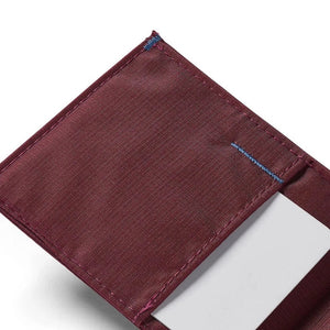 An ALLETT Sport Wallet in RFID-blocking mulberry nylon, along with a pen, sunglasses, candle, seashell, iPhone, and AirPods are neatly displayed on a marble surface.