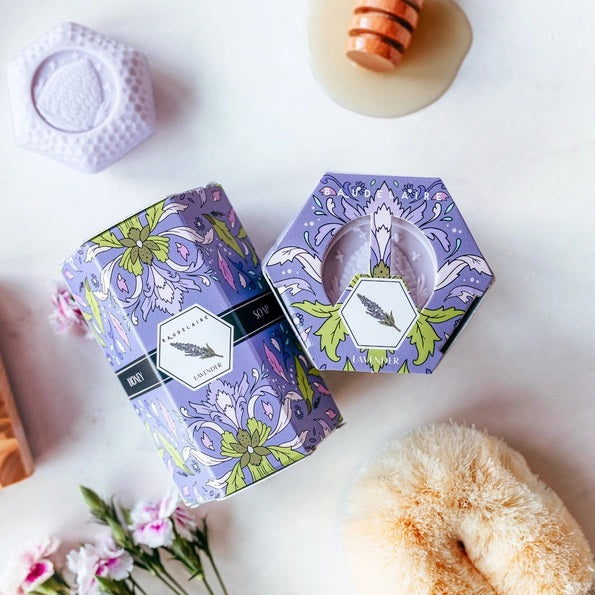 Four lavender soap bars are neatly stacked beside a purple box accented with floral designs and decorative yellow and green details, labeled “BAUDELAIRE - LAVENDER HONEY SOAP 1.4 OZ QUARTET.” From the BAUDELAIRE brand, these luxurious triple-milled soaps create a rich, creamy lather.
