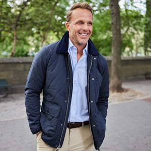A man with light brown hair smiles while standing outside in a park. He wears the COLLARS AND CO - MIDDLEBURG QUILTED FIELD JACKET by COLLARS AND CO, featuring a corduroy collar, layered over a light blue shirt and tan pants for lightweight warmth on a cool day. Trees and a stone wall are visible in the background.