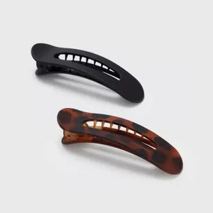 Two sustainable rectangular hair clips, one solid black and the other with a brown and black tortoiseshell pattern, rest on a light pink card. The card emphasizes the recycled materials of "KITSCH - FLAT LAY CLAW CLIP 2 PC," showcasing their style and eco-friendly commitment.