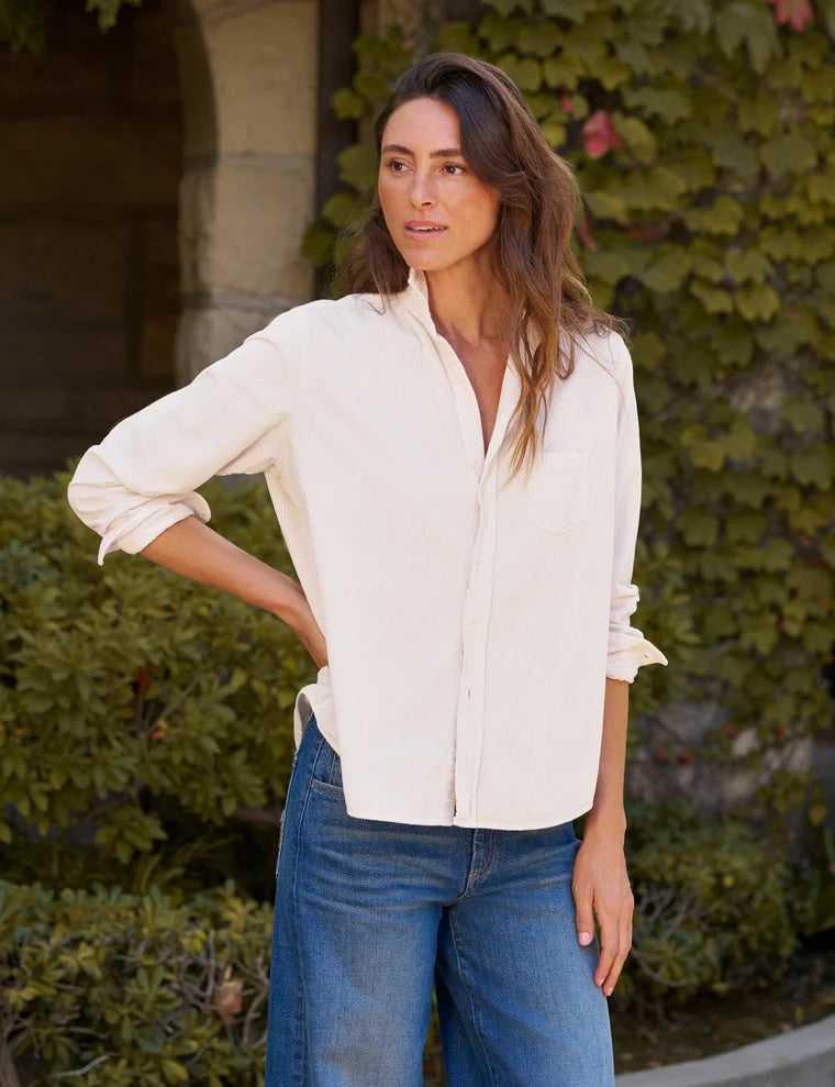 A woman stands outdoors with her hands in her pockets, looking down, wearing the FRANK &amp; EILEEN EILEEN Relaxed Button Up Shirt in Italian Corduroy Vintage White paired with blue jeans. Lush greenery and a stone building create a serene backdrop.