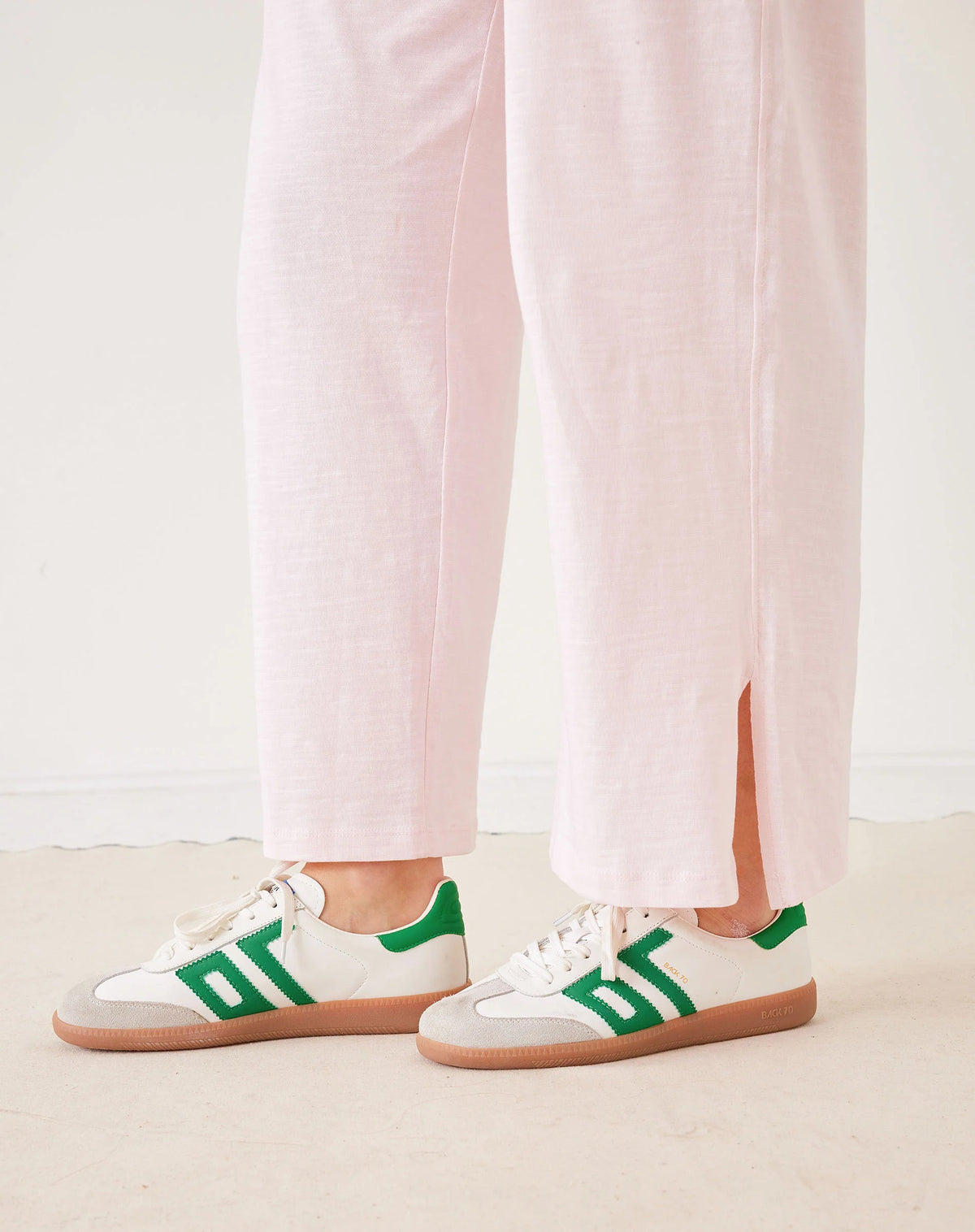A woman stands in a minimal white room wearing the MERSEA TRAVELER TEE PANT, light gray, made from Turkish cotton slub—styled with white sneakers featuring green accents—and smiles softly at the camera.