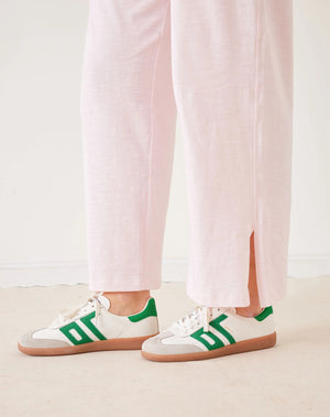 A woman stands in a minimal white room wearing the MERSEA TRAVELER TEE PANT, light gray, made from Turkish cotton slub—styled with white sneakers featuring green accents—and smiles softly at the camera.