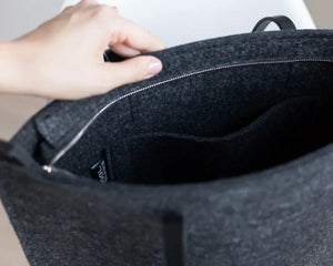 A person in a beige trench coat holds the WOOLBERRY WOOL FELT LONG TOTE BAG with black handles, standing against a plain, light-colored background.