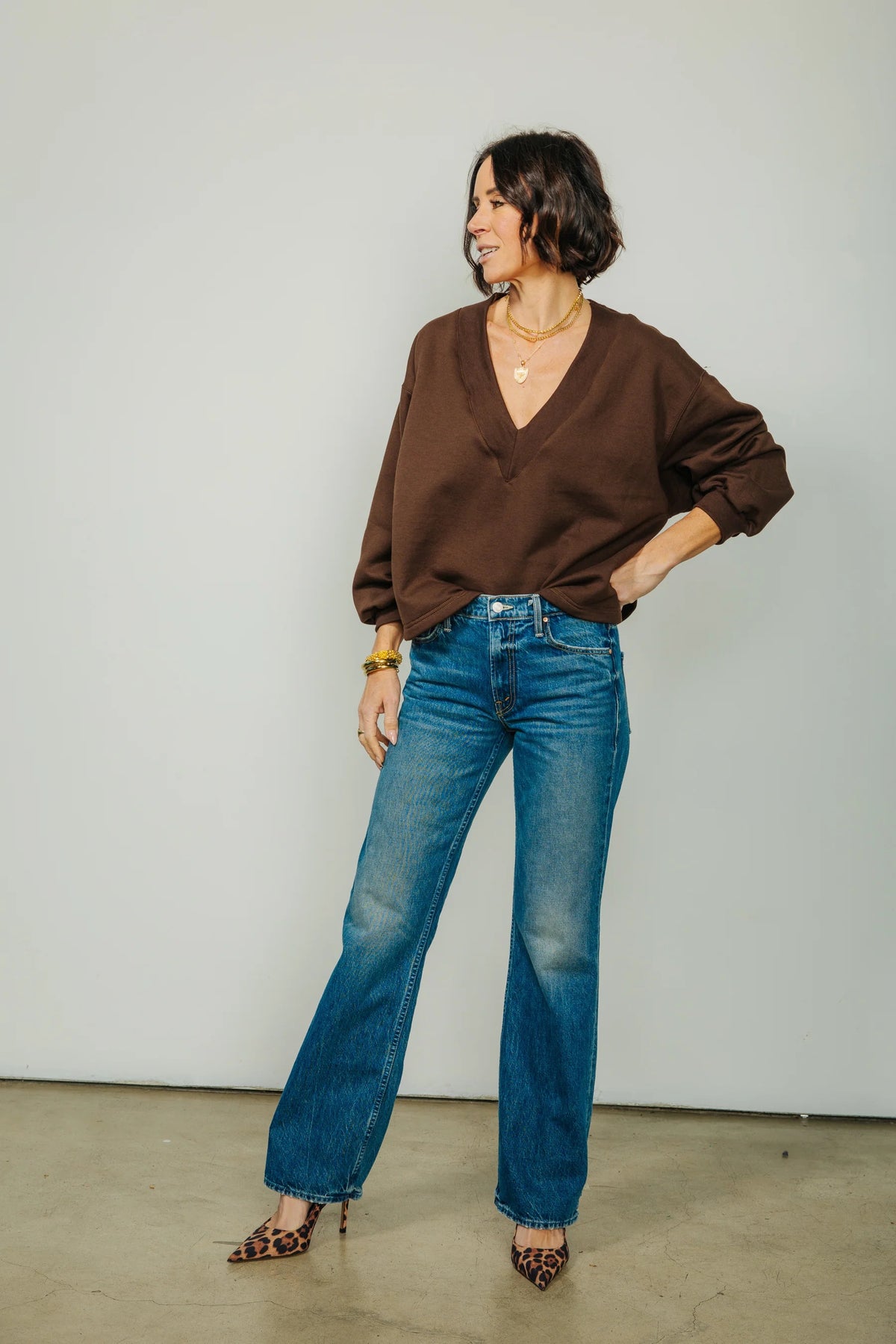 A woman stands indoors, smiling with her hands in her pockets, wearing the KMJ Tucker V Neck Sweater in Chocolate by KMJ—a relaxed fit brown long-sleeve top—paired with blue jeans and leopard print heels. A large window is visible in the background.