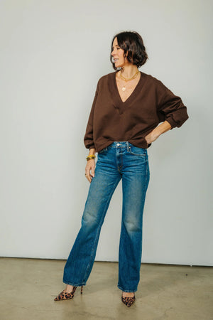 A woman stands indoors, smiling with her hands in her pockets, wearing the KMJ Tucker V Neck Sweater in Chocolate by KMJ—a relaxed fit brown long-sleeve top—paired with blue jeans and leopard print heels. A large window is visible in the background.