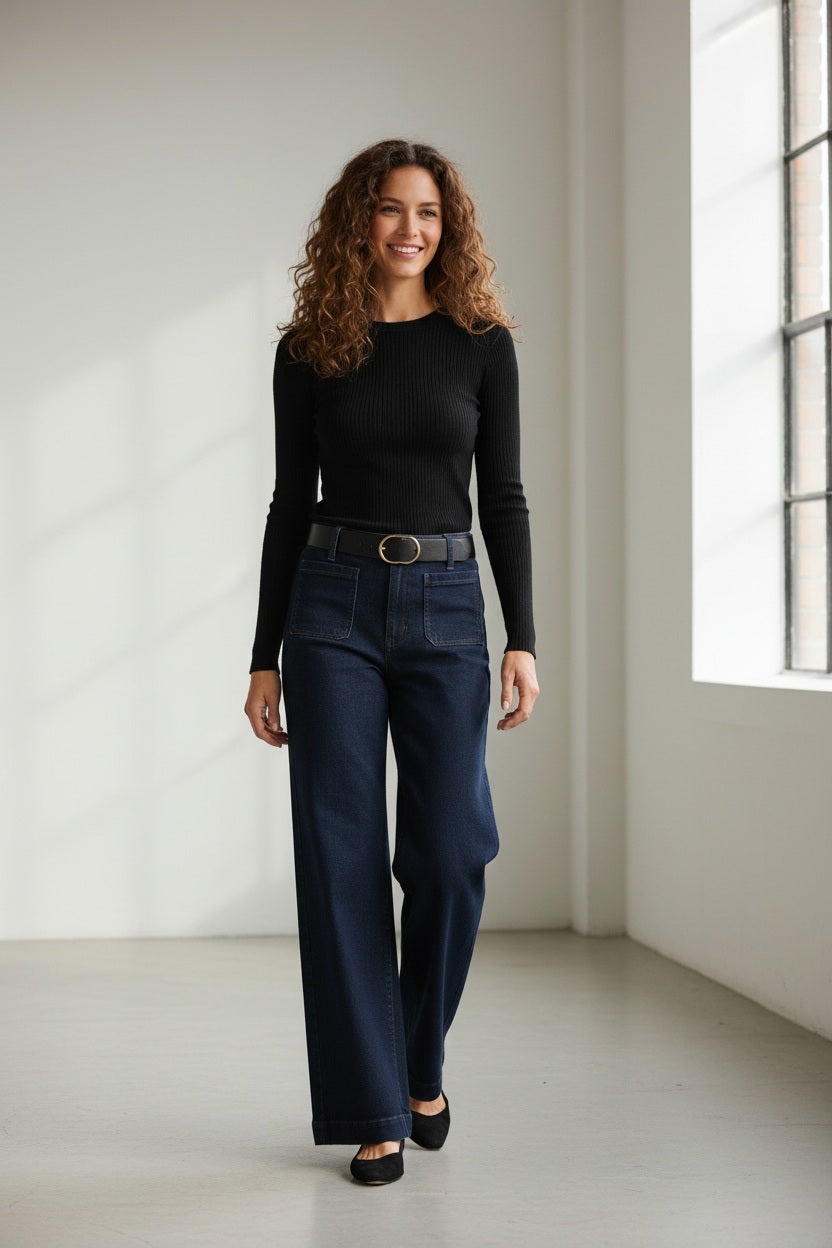 A person wearing MERSEA - SAILOR WIDE LEG JEANS IN RINSE by MERSEA, styled with a black top, black belt, and black heeled sandals, stands with their back to the camera against a plain light background.
