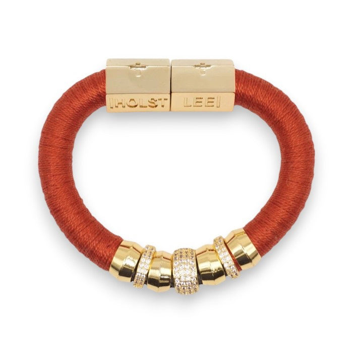 The HOLST + LEE - LUXE CLASSIC BRACELET by HOLST &amp; LEE features an elegant orange cord with gold-plated accents, including a rectangular gold clasp adorned with several gold and diamond-encrusted beads near the clasp.