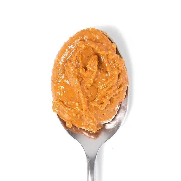 A spoonful of BIG SPOON ROASTERS - SWEET HEAT PEANUT BUTTER against a plain white background. The peanut butter appears creamy with visible small chunks of peanuts, giving it a slightly rough texture. The spoon is metallic and reflects some light, highlighting the spicy essence of this popular condiment from BIG SPOON ROASTERS.