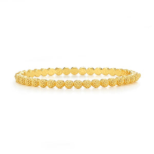 A woman with long blonde hair dons a white off-shoulder top and an 18K gold necklace featuring a pearl pendant. Her accessories include matching earrings and the CAPUCINE DE WULF BERRY ETERNITY PETITE HINGED BANGLE, all set against a backdrop of lush greenery.