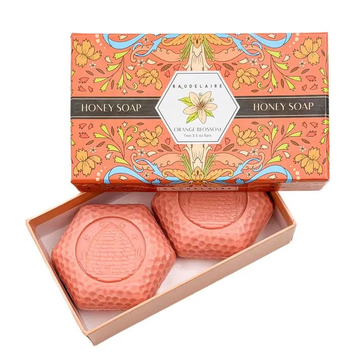 Experience the BAUDELAIRE Orange Blossom Honey Soap 3.5 oz, 2 Bar Gift Box—a beautifully presented set featuring two hexagonal orange soaps nestled in an open decorative box adorned with floral designs and a central bee illustration. This triple-milled soap by BAUDELAIRE offers a luxurious lather and a delicate orange blossom honey fragrance, making it an ideal indulgence for yourself or a thoughtful gift for someone special.