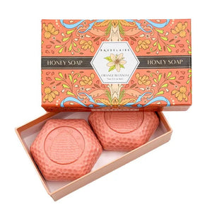 Experience the BAUDELAIRE Orange Blossom Honey Soap 3.5 oz, 2 Bar Gift Box—a beautifully presented set featuring two hexagonal orange soaps nestled in an open decorative box adorned with floral designs and a central bee illustration. This triple-milled soap by BAUDELAIRE offers a luxurious lather and a delicate orange blossom honey fragrance, making it an ideal indulgence for yourself or a thoughtful gift for someone special.