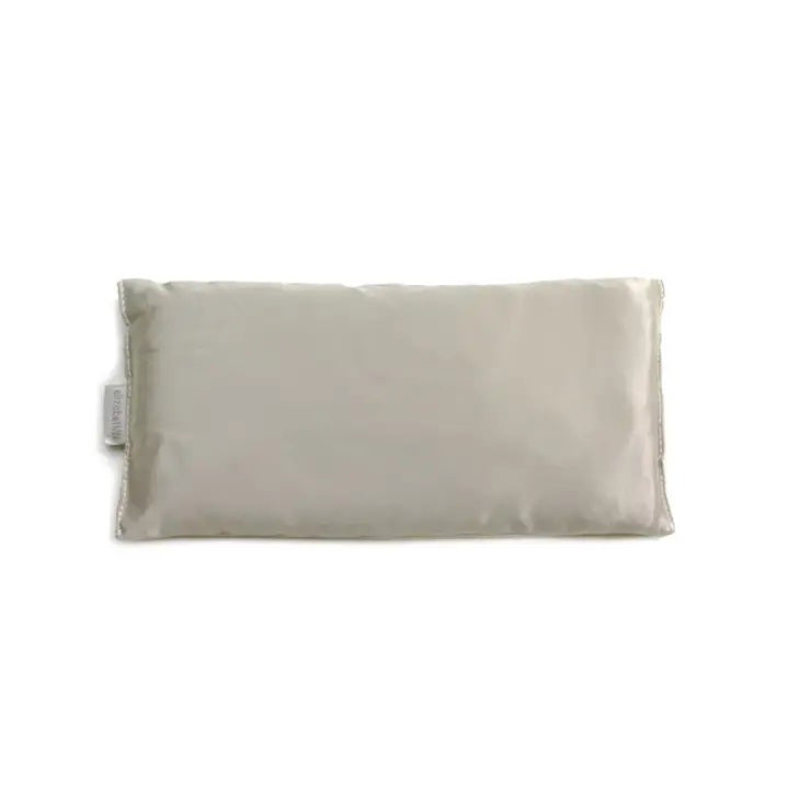 The SILK EYE PILLOW - SILVER by E WIGHTMAN & CO features a rectangular, light gray design with a smooth texture and a small fabric tag on the left side. This simple and minimalist eye pillow, filled with flaxseed for enhanced relaxation, is showcased on a white background.
