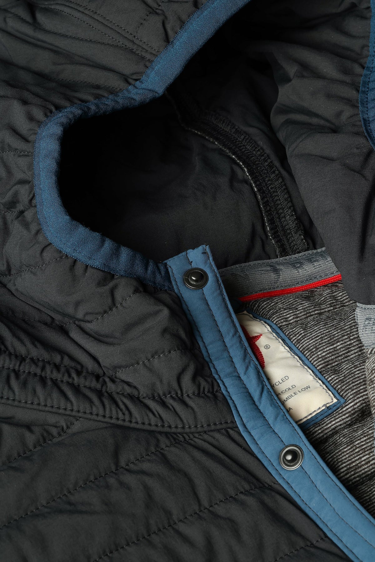 Wearing the RELWEN - WINDZIP HOOD IN BLACK FADE, a black water-resistant jacket with recycled polyester insulation from RELWEN, a person stands on a snowy path in blue pants, covering their face with their hand. Snow falls gently as snow-laden trees fill the background.