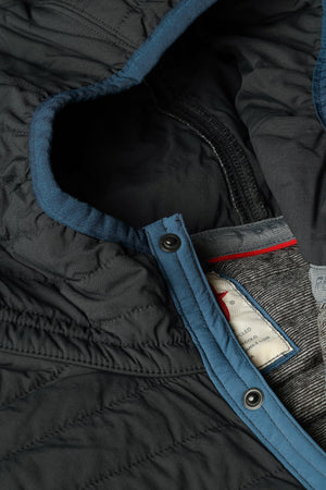 Wearing the RELWEN - WINDZIP HOOD IN BLACK FADE, a black water-resistant jacket with recycled polyester insulation from RELWEN, a person stands on a snowy path in blue pants, covering their face with their hand. Snow falls gently as snow-laden trees fill the background.