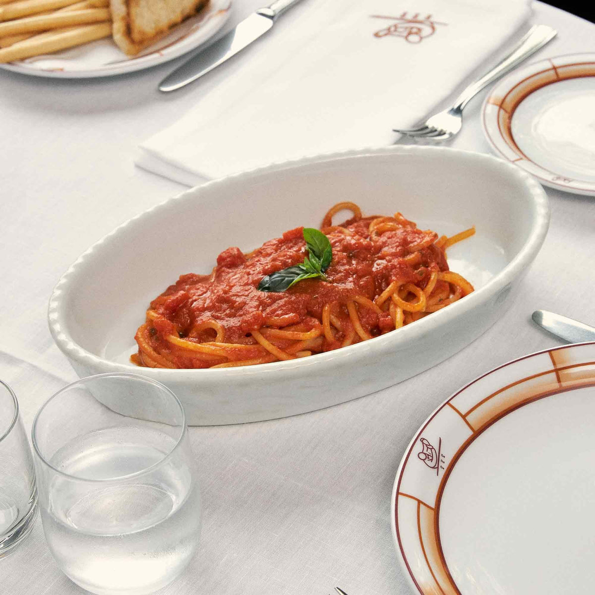 A white oval dish of Atalanta Corporation’s CIPRIANI – ORGANIC SPAGHETTI—made from premium organic durum wheat semolina—is topped with vibrant red tomato sauce and a fresh basil leaf, set atop a white tablecloth and surrounded by plates, cutlery, napkins, and glasses of water.