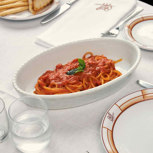 A white oval dish of Atalanta Corporation’s CIPRIANI – ORGANIC SPAGHETTI—made from premium organic durum wheat semolina—is topped with vibrant red tomato sauce and a fresh basil leaf, set atop a white tablecloth and surrounded by plates, cutlery, napkins, and glasses of water.