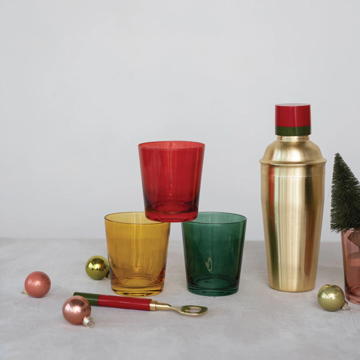Three colorful CREATIVE COOP LOW BALL GLASSWARE pieces (red, yellow, green) are stacked next to a gold cocktail shaker with a red top, decorative ornaments, and a bar spoon, with a small tree in the background.