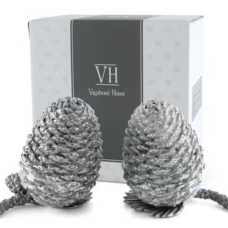 Two detailed silver pine cones stand upright on a white background, resembling the VAGABOND HOUSE - PEWTER PINE CONESALT AND PEPPER SET by VAGABOND HOUSE, with small pine needle segments visible at their bases.