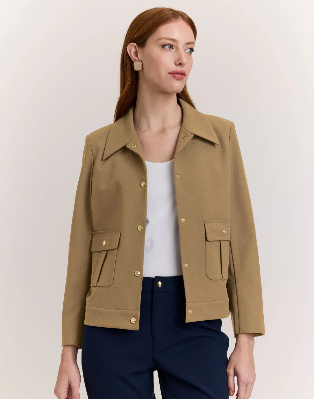 A woman with long red hair wears the HINSON WU HANNAH Long Sleeve Traveler’s Twill Jacket in tan over a white top and navy pants, standing against a plain light background and looking slightly to the side.