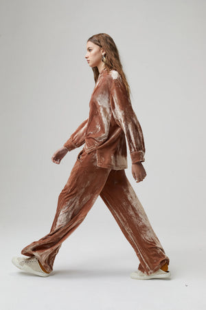 A person with long wavy hair stands against a plain background wearing a mustard yellow, pure silk velvet long-sleeve top and matching, high-waisted LANHTROPY - THE LUNA VELVET PANT by LANHTROPY. They are accessorized with layered necklaces, a brown belt, and flat sandals.