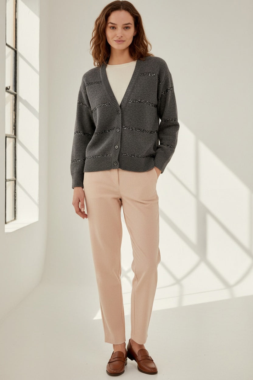 A woman with wavy brown hair models the ANNECLAIRE - PAILETTES STRIPE CARDIGAN, a light grey cotton piece with subtle horizontal stripes, paired with beige pants against a plain background.