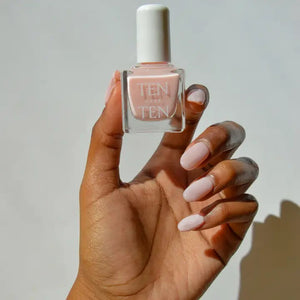 A glass bottle of TENOVERTEN - NAIL POLISH from the brand TEN OVER TEN, featuring a white cap and a dusty pink nail color. The front label displays "TEN OVER TEN" in white uppercase letters. This vegan, eight-free formula ensures both beauty and peace of mind. The background is plain and light-colored.