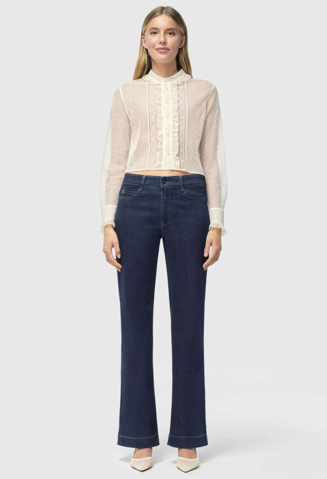 A person with light brown hair stands against a plain background, wearing a sheer, ruffled, long-sleeved white blouse and MAC JEANS - DREAM WIDE AUTHENTIC by MAC in dark blue. They&#39;re paired with comfortable white shoes.