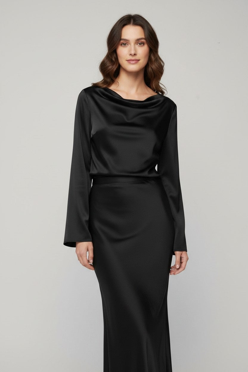 A woman stands against a light gray background, wearing the MILVA MI Satin Blouse in Black by MILVA. The blouse features long sleeves and a draped cowl neckline, and she pairs it effortlessly with wavy brown hair and a slight smile, exuding relaxed sophistication with its elegant silhouette.
