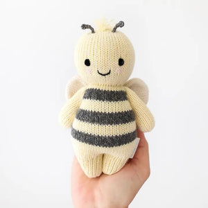 A baby lies on a cozy white blanket, raising both arms. The baby is dressed in a peach outfit and a headband with three pom-poms. Next to the baby is the CUDDLE + KIND - HANDMADE BABY BEE by CUDDLE & KIND, a fair trade, hand-knit toy with black and white stripes—a perfect addition to any collection of baby shower gifts.