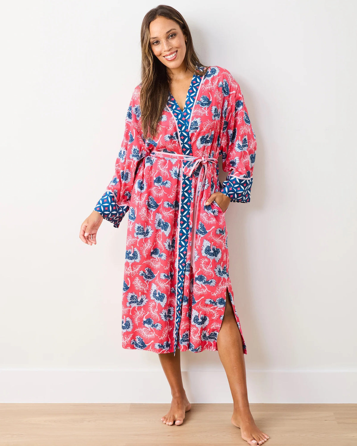 A smiling person, barefoot on wooden flooring against a white wall, wears the vibrant PRINTFRESH - SATIN ROBE OSTRICH DANCE. This eco satin robe has blue and pink floral patterns, a geometric trim, and a waist tie for comfort and style.