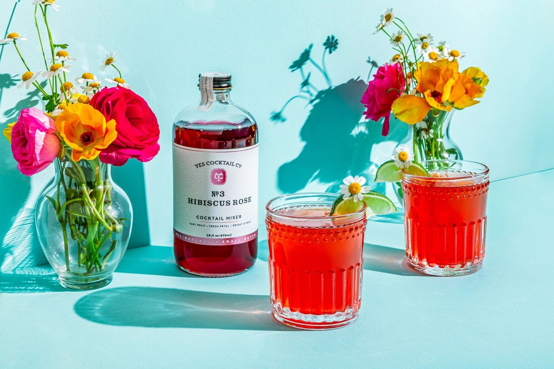 A transparent glass bottle holds a rich, dark red liquid labeled "YES COCKTAIL CO - Hibiscus Rose Cocktail Mixer." Handcrafted in California and available in a 16 fl oz (473ml) size, the white-and-red-toned label indicates its perfect suitability for creating cocktails or an exotic floral spritzer.