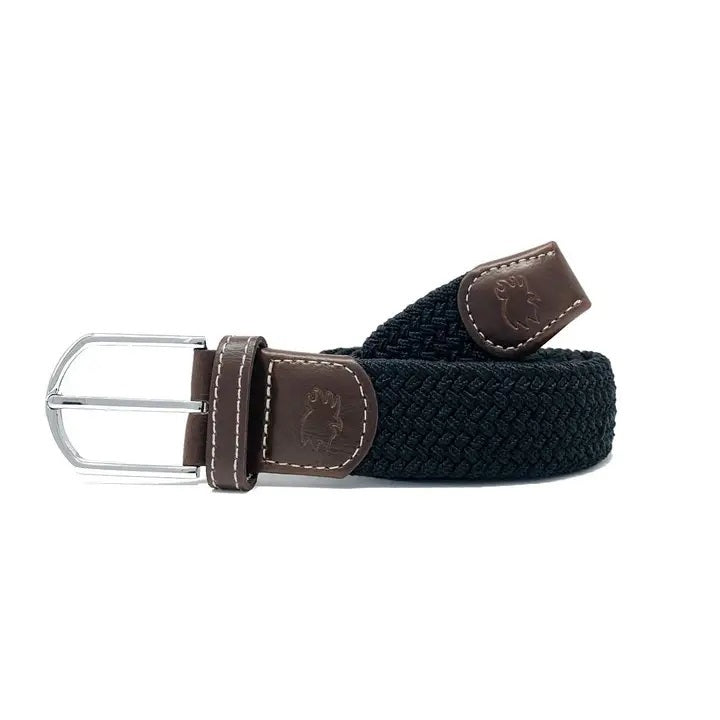 Outdoors, a person with hands in pockets wears a navy blue shirt, light-colored pants, and the ROOSTAS - BETHPAGE WOVEN STRETCH BELT by ROOSTAS featuring brown leather accents.
