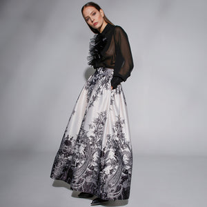 A woman stands against a plain gray backdrop wearing a sheer black blouse with ruffle details at the chest and MILVA's LONG MAXI SKIRT, featuring a detailed black-and-white pattern. The high-waisted, floor-length skirt, with welt pockets and a side closure with concealed zip, cinches at her gathered waist. She wears black shoes.