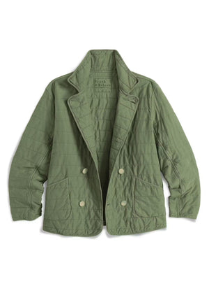 A woman with short, wavy dark hair stands indoors in a bright room with large windows, wearing the FRANK & EILEEN Quilted Belfast Jacket in Vintage Green over a white turtleneck, brown pants, and dark loafers. The light green jacket is made from Italian peached twill.