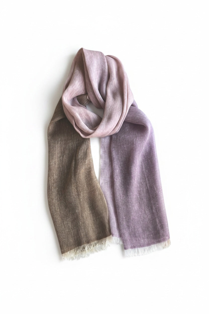 The BAJRA Hand Dyed Linen Blend Scarf in Plinth Brown Ombre showcases a color gradient from pink to purple to brown, elegantly looped against a white backdrop. This handmade piece features fringed edges and embodies the warmth and craftsmanship of Nepal.