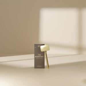 A gold THE ASSISTANT KEY TOOL by AKT LONDON and its beige, rectangular box are displayed on a smooth surface with soft sunlight and shadows, showcasing this elegant, plastic-free addition to your routine.