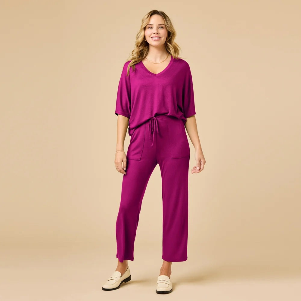 A woman wears a SOFTIES - DREAM RELAXED V-NECK WITH CAPRI LOUNGE SET in magenta, featuring a V-neck top with elbow-length sleeves and matching cropped drawstring pants, paired with cream-colored loafers.