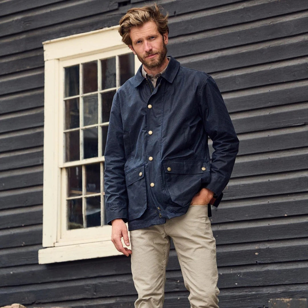 The TOM BECKBE Piedmont Jacket in Navy features waxed cotton, long sleeves, a collar, snap button closures, two large front pockets, and a contrasting interior lining. This weatherproof jacket combines classic style with durability.