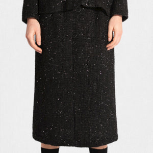 A person is wearing a stylish, textured black MEIMEIJ Lurex Tweed suit, featuring a tailored jacket with fringe details and the MEIMEIJ - LUREX TWEED SKIRT. Underneath the jacket is a white T-shirt with bold black text. The outfit is paired with black socks and black shoes.