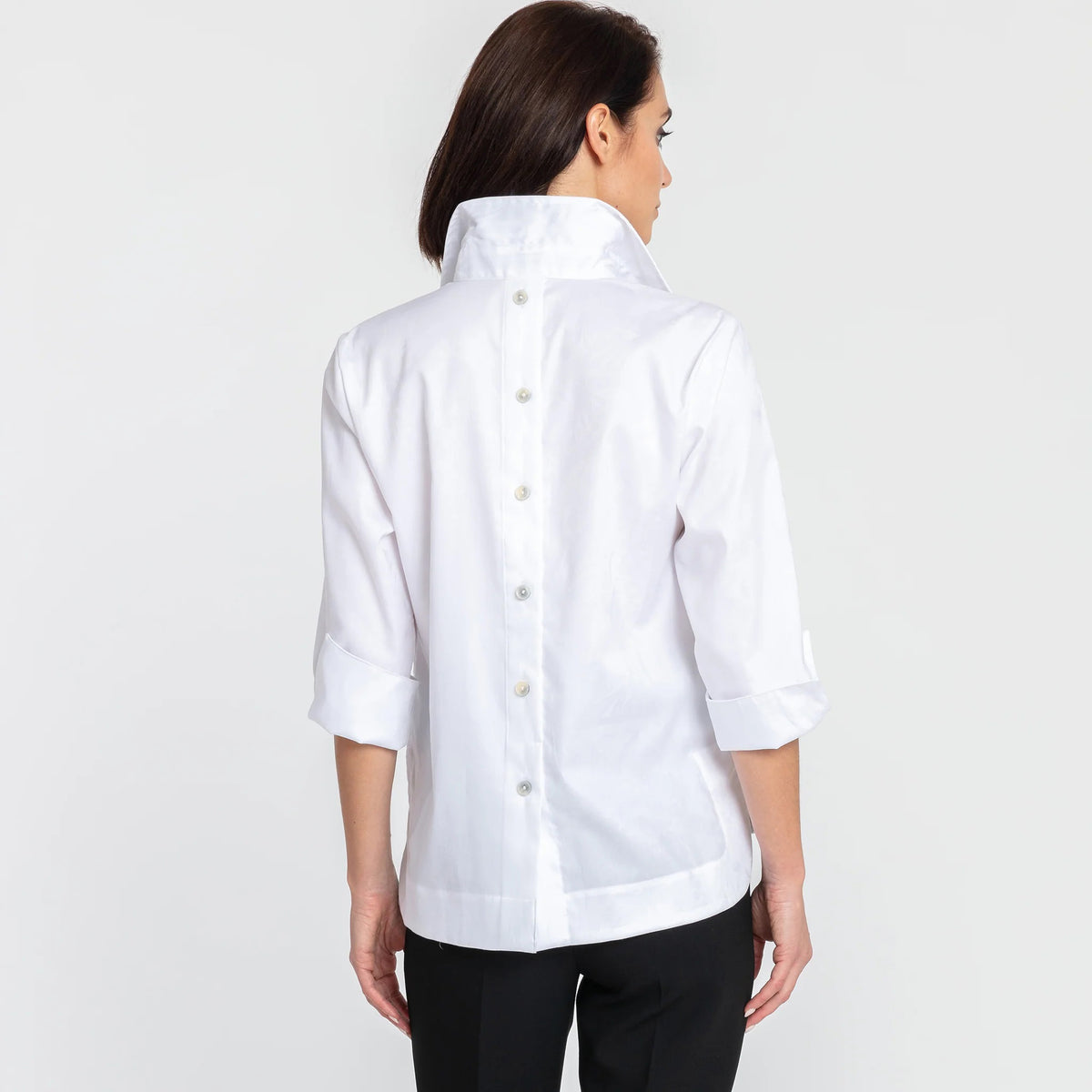 A woman with short dark hair models the HINSON WU - AILEEN 3/4 SLEEVE POLISHED COTTON SHIRT in navy, featuring a collared neckline, stretch fabric, and three buttons. She pairs it with white pants and poses against a plain light gray background.