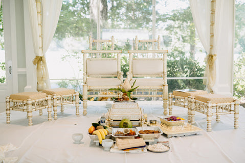 Ornate gold chairs and stools are arranged in a bright room with large windows. In front, ritual items and a variety of fruits and offerings are placed on a white cloth, suggesting a ceremonial or religious setup.