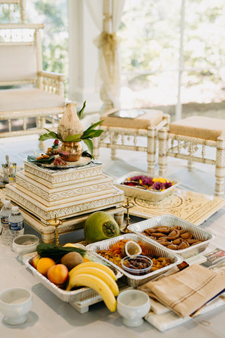 A traditional Hindu puja setup with a decorative altar, fruit offerings, sweets, flowers, and ritual items arranged on a table, surrounded by ornate chairs in a bright, airy setting.
