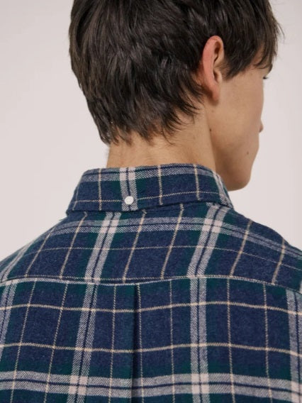 A young person with dark hair, wearing the HARTFORD - PITT CHECKED FLANNEL SHIRT IN BLUE AND GREEN by HARTFORD, looks at the camera while adjusting the shirt collar with one hand against a plain light background.