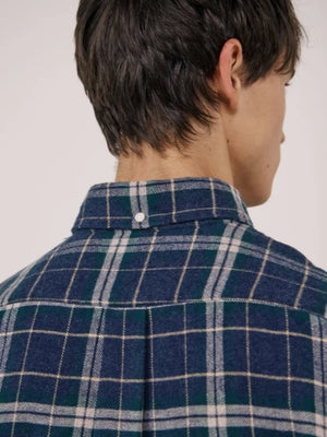 A young person with dark hair, wearing the HARTFORD - PITT CHECKED FLANNEL SHIRT IN BLUE AND GREEN by HARTFORD, looks at the camera while adjusting the shirt collar with one hand against a plain light background.