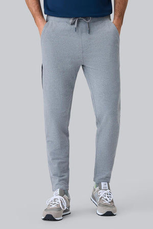 A person wearing MACK WELDON ACE Sweatpants in Ice Storm Heather, a dark blue shirt, and gray sneakers with white soles stands on a light gray background, shown from the side with one arm relaxed.