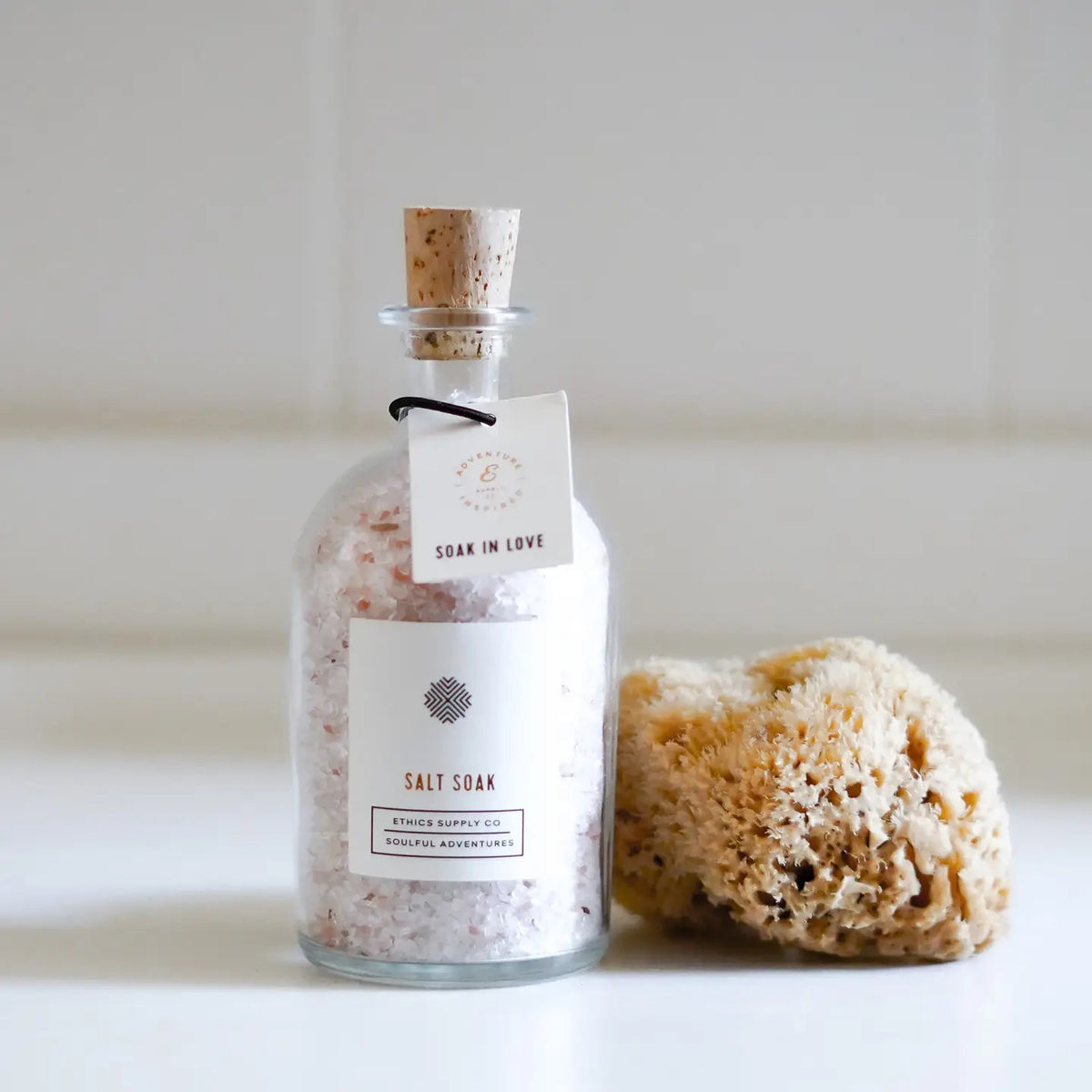 A glass bottle of cruelty-free, coarse pink "Soak in Love" bath salt by Ethics Supply Co, sealed with a cork stopper, rests on a white surface. Nearby, a natural sea sponge awaits, promising an indulgent escape with each use.